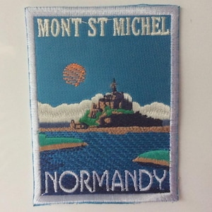 May include: Embroidered patch featuring the words "MONT ST MICHEL" and "NORMANDY" above and below a depiction of the island's abbey. The design includes a blue sky with an orange sun, the abbey on a rocky island, and the surrounding sea.