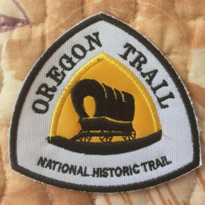 May include: A triangular patch with the words "OREGON TRAIL" and "NATIONAL HISTORIC TRAIL" in black. The centre has a yellow background with a black illustration of a covered wagon.