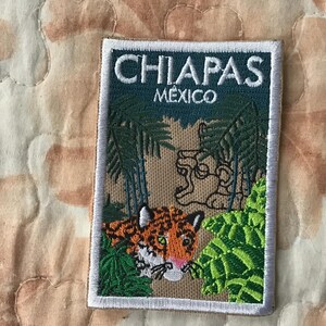 Puede incluir: Un parche rectangular bordado con el texto CHIAPAS MEXICO en la parte superior. El diseño muestra una escena de selva con palmeras verdes, una talla de piedra y un tigre en primer plano con detalles de pelaje naranja, negro y blanco.