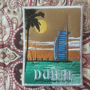 Puede incluir: Parche bordado con una escena de Dubái con una palmera, un sol amarillo y un edificio azul y blanco distintivo. La palabra "DUBAI" está bordada en blanco sobre un fondo verde, con "UNITED ARAB EMIRATES" debajo.
