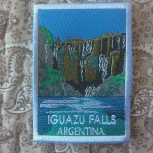 以下が含まれることがあります： アルゼンチンのイグアスの滝を描いた刺繍パッチ。デザインには、青い空、緑と茶色の崖、青い川に流れ込む滝が含まれています。「IGUAZU FALLS ARGENTINA」の文字が白で表示されています。
