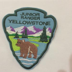 May include: Embroidered patch with a light blue background featuring the text "JUNIOR RANGER YELLOWSTONE". The patch depicts a brown bear near a river, with mountains and trees in the background.