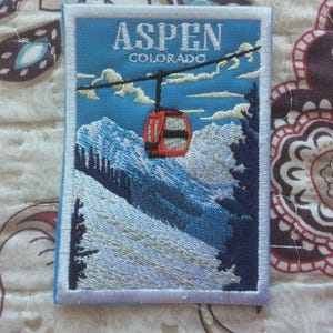 May include: Embroidered patch featuring a ski lift over snow-covered mountains, with the text "ASPEN COLORADO" in white. The patch has a blue background and a white border, depicting a scenic mountain landscape.