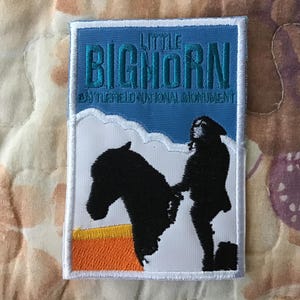 May include: A rectangular embroidered patch featuring a blue sky background with the text LITTLE BIGHORN BATTLEFIELD NATIONAL MONUMENT in teal. The lower portion shows a white landscape with a black silhouette of a person and a horse.