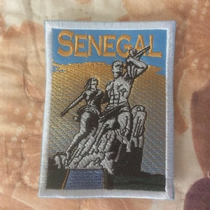 Könnte beinhalten: Gestickter Aufnäher mit dem Wort "SENEGAL" und einer Darstellung eines Denkmals mit zwei Figuren. Der Aufnäher hat einen blauen Rand und einen gold-blauen Hintergrund.