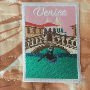 May include: Embroidered patch featuring a Venice scene with a bridge, gondola, and buildings. The patch has a pink background with the word "Venice" in white script. The patch is bordered with white stitching.