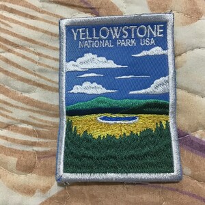 May include: A rectangular embroidered patch featuring a landscape of Yellowstone National Park USA. The design includes a blue sky with white clouds, green mountains, a yellow meadow, and a small blue lake, all bordered by a silver-grey stitched edge.