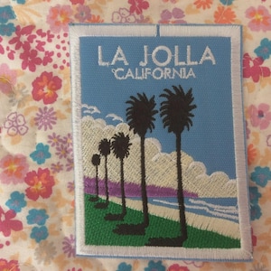 Puede incluir: Parche bordado con una escena costera con palmeras y el texto "LA JOLLA CALIFORNIA" sobre un cielo azul. El diseño incluye una zona de césped verde, una playa de arena y nubes blancas.