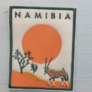 Könnte beinhalten: Gestickter Aufnäher mit einer namibischen Landschaft. Das Design umfasst eine orangefarbene Sonne, Wüstenlandschaft und eine Oryx. Das Wort "NAMIBIA" ist oben in Großbuchstaben aufgestickt. Der Aufnäher hat einen grünen Rand.