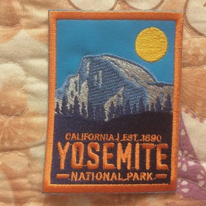 May include: Embroidered patch featuring a mountain landscape under a yellow sun against a blue sky. The patch reads "YOSEMITE NATIONAL PARK" with "CALIFORNIA | EST. 1890" above. The patch has an orange border.