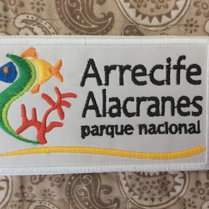 May include: Embroidered patch featuring a colorful fish, coral, and the text "Arrecife Alacranes parque nacional." The design includes a yellow line at the bottom and is set against a light gray background with a white border.