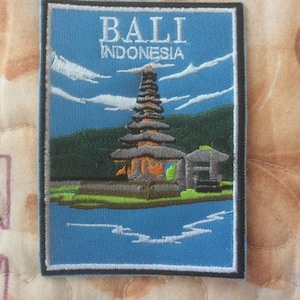 Puede incluir: Parche bordado con una escena de un templo balinés con el texto "BALI INDONESIA". El parche tiene un fondo azul con nubes blancas, un templo escalonado y una pequeña estructura. El parche está bordeado en negro.
