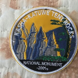 Op de afbeelding: Een ronde geborduurde patch met een gele luchtachtergrond en grijze en blauwe rotsformaties. De tekst KASHA-KATUWE TENT ROCKS staat in een boog bovenaan, met NATIONAL MONUMENT en 2001 in het wit op een zwarte basis aan de onderkant.