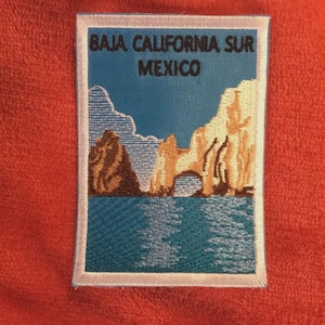 May include: Embroidered patch depicting a coastal scene from Baja California Sur, Mexico. The design shows a turquoise sea, tan rock formations, and the text "BAJA CALIFORNIA SUR MEXICO" in black, within a white border.