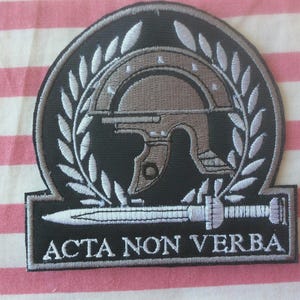 May include: Embroidered patch with a Roman helmet, sword, and laurel wreath design. The patch is black with grey and white details, and the text "ACTA NON VERBA" is displayed in white.