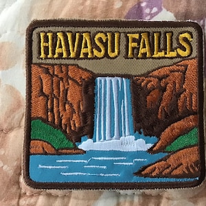 May include: A square embroidered patch featuring a landscape of Havasu Falls. The design includes a light blue waterfall cascading between brown canyon walls, green foliage at the base, and a light blue pool. The text HAVASU FALLS is in yellow.