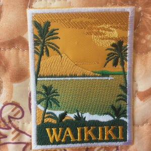 Puede incluir: Parche bordado con una escena tropical con palmeras, una montaña y la palabra "WAIKIKI" en letras doradas. El parche tiene un borde blanco y una combinación de colores amarillo y verde, que evoca una puesta de sol sobre el océano.
