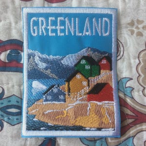 Puede incluir: Parche bordado con un paisaje de Groenlandia. El parche tiene un fondo azul con la palabra "GREENLAND" en blanco. El diseño incluye montañas, casas coloridas y una masa de agua.