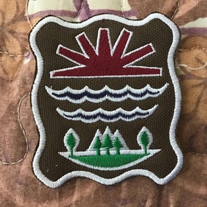 Pode incluir: Patch bordado com fundo castanho e borda branca. O design apresenta um sol vermelho, ondas azuis, árvores verdes e um contorno branco. Este patch é provavelmente um emblema decorativo.