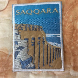 May include: Embroidered patch with the word "SAQQARA" above a landscape scene. The patch features a blue sky, tan structures, and a white border. The design is detailed with various architectural elements.