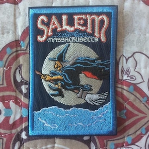 May include: Embroidered patch featuring a witch on a broomstick flying in front of a full moon. The patch has the word "Salem" arched across the top, with "Massachusetts" below. The design is framed in blue.