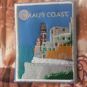 May include: Embroidered patch featuring a coastal scene with the text "AMALFI COAST." The design includes a blue sky and sea, a detailed depiction of buildings, and a tower. The patch is bordered with white stitching.