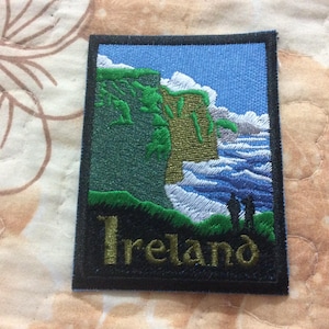 Op de afbeelding: Geborduurde patch met een schilderachtig uitzicht op de Ierse kust. Het ontwerp toont groene kliffen, een blauwe oceaan en een blauwe lucht met witte wolken. Het woord "Ireland" is goudkleurig geborduurd.