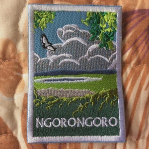 May include: Embroidered patch depicting a landscape with a butterfly, clouds, and foliage. The patch shows a blue sky, green grass, and the word "NGORONGORO" in white. The patch is rectangular with a white border.