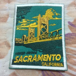 以下が含まれることがあります： カリフォルニア州サクラメントのタワーブリッジを描いた刺繍パッチ。デザインは、橋を黄色で、ティール色の背景に、都市名と州名を下に表示しています。パッチは白い縁取りがあります。