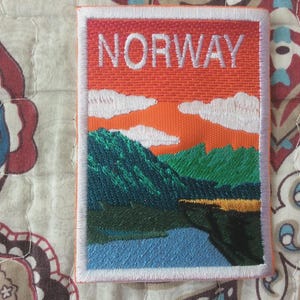 May include: Embroidered patch featuring a landscape scene with the word "NORWAY" in white block letters. The patch depicts a mountain range, a body of water, and a colorful sky with clouds. The patch is bordered with white stitching.
