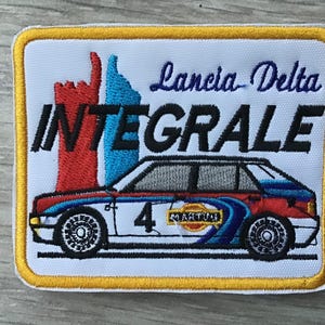 May include: A rectangular embroidered patch with a yellow border on a white background. It features the text Lancia Delta INTEGRALE in black and blue, a red and blue abstract graphic, and a white rally car with the number 4 and Martini logo.