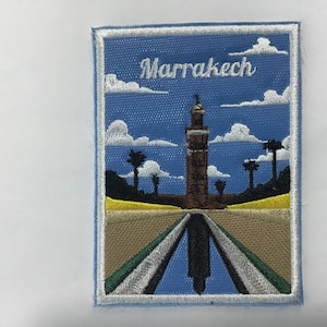 Pode incluir: Patch bordado com uma cena de Marraquexe, com um alto minarete, palmeiras e o nome da cidade em escrita branca sobre um céu azul com nuvens. O patch tem uma borda azul clara e um design detalhado e colorido.