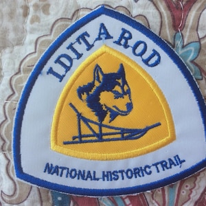 May include: Embroidered patch with a blue and white border, featuring the text "Iditarod National Historic Trail." The patch has a yellow center with a blue husky dog and sled design.