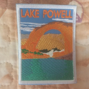 May include: Embroidered patch featuring a scenic view of Lake Powell. The patch displays the words "LAKE POWELL" above an orange rock formation and blue water. The patch has a white border.