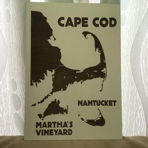 May include: A rectangular wooden sign featuring a dark brown laser-cut map of Cape Cod, Nantucket, and Martha's Vineyard on a light cream background. The text CAPE COD, NANTUCKET, and MARTHA'S VINEYARD is printed in a bold, dark brown sans-serif font.