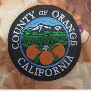 Può includere: Toppa circolare nera ricamata con le parole "COUNTY OF ORANGE CALIFORNIA" in bianco. La toppa presenta un cielo blu e una scena di montagna sopra campi verdi e tre arance arancioni.