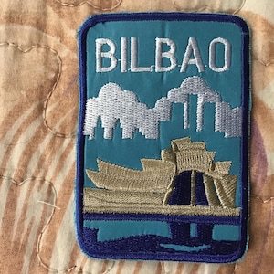 Op de afbeelding: Een rechthoekige geborduurde patch met het woord BILBAO in witte hoofdletters aan de bovenkant. Onder de tekst bevindt zich een zilvergrijze skyline boven een goudkleurige architecturale structuur, alles op een lichtblauwe achtergrond.