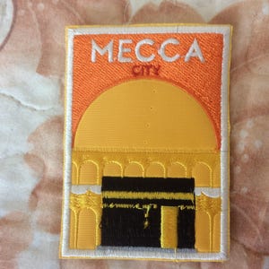 Puede incluir: Parche bordado con la Kaaba en La Meca. El parche tiene forma rectangular, con un fondo naranja y las palabras "MECCA CITY" en blanco. La Kaaba está representada en negro y dorado.