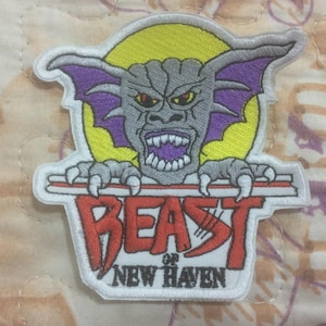 Könnte beinhalten: Gestickter Aufnäher mit einem grauen Monster mit lila fledermausartigen Flügeln und scharfen Zähnen, das hinter einem rot-weißen Banner hervorkommt. Der Text "BEAST of NEW HAVEN" ist darunter abgebildet.