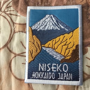 May include: Embroidered patch featuring a mountain landscape with a river. The patch has a blue background with a white-capped mountain and yellow and brown terrain. Text at the bottom reads "NISEKO HOKKAIDO JAPAN."
