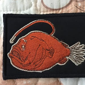 Könnte beinhalten: Ein rechteckiger, schwarzer Stickaufnäher mit einem detaillierten orangefarbenen Anglerfisch, der einen markanten leuchtenden Köder am Kopf trägt. Der Fisch hat scharfe Zähne, einen strukturierten Körper und eine helle, fächerförmige Schwanzflosse.