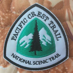 May include: A white, green, and black embroidered patch with the text "PACIFIC CREST TRAIL NATIONAL SCENIC TRAIL". The patch features a green triangle with a black tree and white mountain range design.