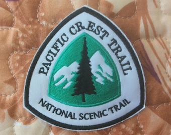 Może przedstawiać: Haftowana naszywka w kolorze białym, zielonym i czarnym z napisem "PACIFIC CREST TRAIL NATIONAL SCENIC TRAIL". Naszywka ma zielony trójkąt z czarnym drzewem i białym wzorem gór.
