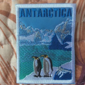 May include: Embroidered patch featuring the word "ANTARCTICA" in blue above a scenic depiction of the Antarctic landscape. Two penguins stand on a snowy shore, with mountains and water in the background.