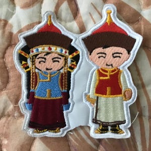 Op de afbeelding: Een geborduurde patch met twee cartoonfiguren in traditionele kleding. De linkerfiguur draagt een blauw vest, een donkerrode rok en een puntige rode muts. De rechterfiguur draagt een rood vest en een lichtgekleurd gewaad met een rode muts.