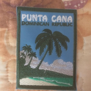 Può includere: Toppa ricamata raffigurante una scena tropicale con palme, spiaggia e onde dell'oceano. La toppa presenta la scritta "PUNTA CANA DOMINICAN REPUBLIC" in bianco su sfondo blu, incorniciata da un bordo verde.