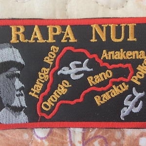 May include: Embroidered patch with the text "RAPA NUI" in gold letters. The patch features a map of Easter Island with place names and a Moai statue. The patch has a black background with a red border.