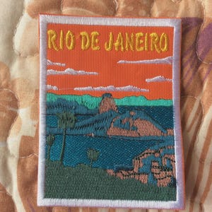 Könnte beinhalten: Gestickter Aufnäher mit einer Landschaft von Rio de Janeiro und dem Text "RIO DE JANEIRO" in Gelb. Das Design zeigt einen orangefarbenen Himmel, blaues Wasser, grüne Hügel und Palmen, alles umrahmt von einem weißen Rand.