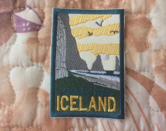 May include: Embroidered patch featuring a landscape scene with a mountain, water, and sky. The patch has a teal border and the word "ICELAND" in gold lettering. The background is a quilted fabric with floral and heart patterns.