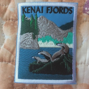 Puede incluir: Parche bordado con una vista panorámica de los fiordos de Kenai. El parche representa montañas, árboles y focas en un paisaje azul y verde. El texto "KENAI FJORDS" está bordado en negro en la parte superior.